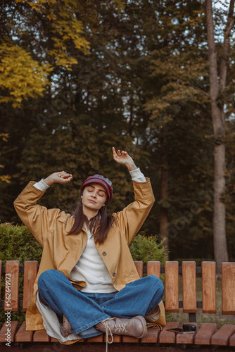 Carefree woman with closed eyes enjoying music in earphones while sitting on bench in autumn park 