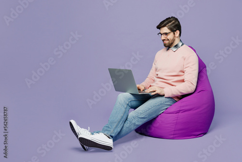 Print op canvas Full body fun young caucasian IT man he wear casual clothes pink sweater glasses sit in bag chair hold use work on laptop pc computer isolated on plain pastel light purple background studio portrait