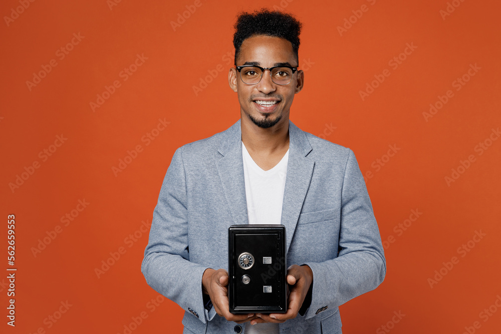Young fun employee business man corporate lawyer wear classic formal grey suit shirt glasses work in office hold metal bank safe for money accumulation isolated on plain red orange background studio. Young fun employee business man corporate lawyer wear classic formal grey suit shirt glasses work in office hold metal bank safe for money accumulation isolated on plain red orange background studio.