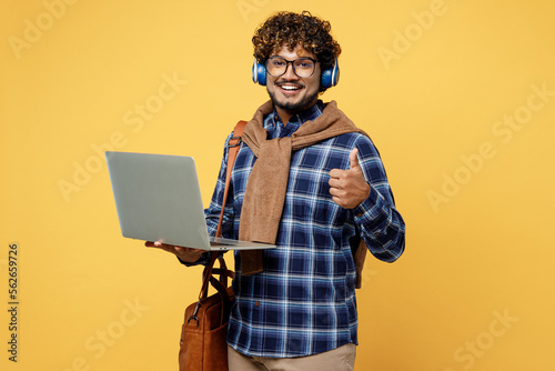 Young teen Indian boy IT student wear casual clothes shirt glasses bag headphones use laptop pc computer show thumb up isolated on plain yellow color background High school university college concept