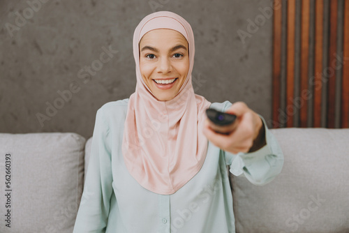 Young muslim woman wears hijab casual clothes watch tv movie film sits on sofa couch stay at home flat rest relax spend free spare time in living room indoor. People middle eastern uae islam concept.
