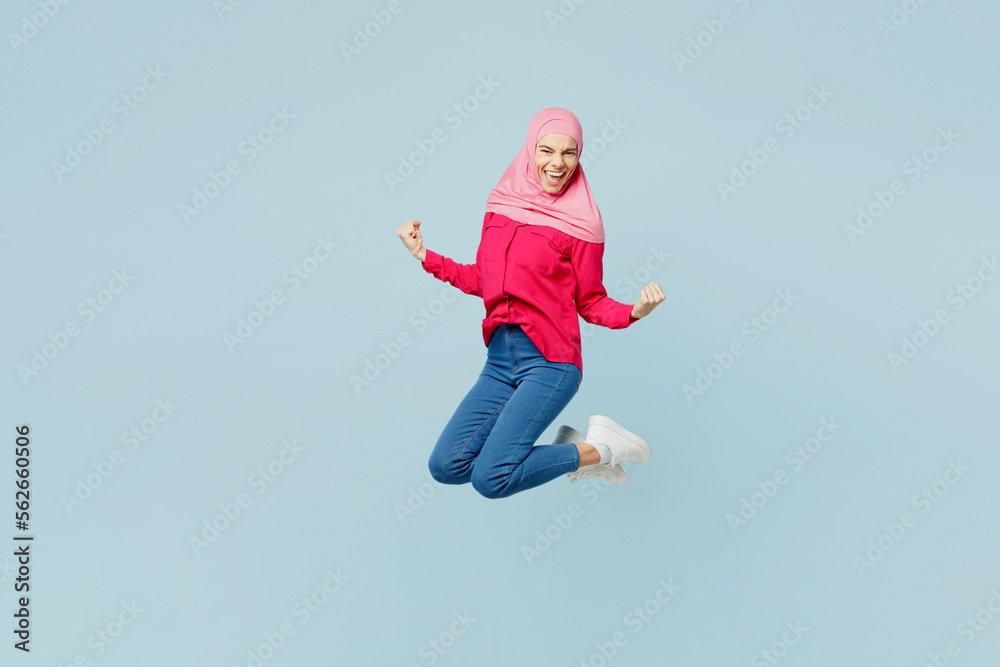 Full body side view young arabian asian muslim woman wearing pink abaya hijab jump high do winner gesture isolated on plain pastel light blue cyan background studio portrait People uae islam concept. Full body side view young arabian asian muslim woman wearing pink abaya hijab jump high do winner gesture isolated on plain pastel light blue cyan background studio portrait People uae islam concept.