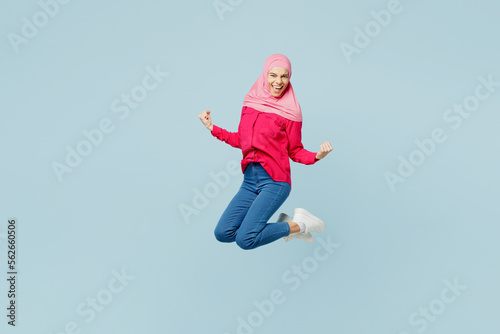 Full body side view young arabian asian muslim woman wearing pink abaya hijab jump high do winner gesture isolated on plain pastel light blue cyan background studio portrait People uae islam concept.