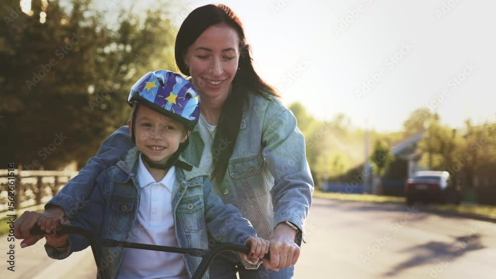 Happy family walk on street the city mother teaches her son child to ride a bike and run nearby. Slow motion woman helps keep balance for safe activity. Sports walk in nature. Healthy lifestyle.