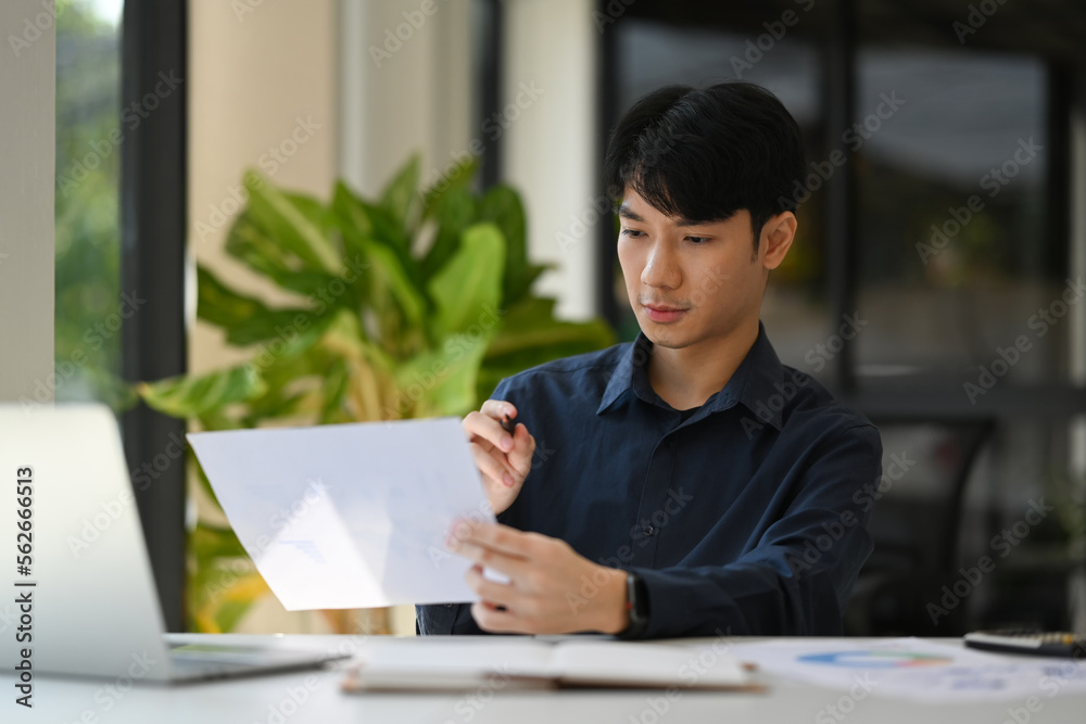 Focused asian male investor sitting front of laptop computer at his workplace and reading financial document Focused asian male investor sitting front of laptop computer at his workplace and reading financial document