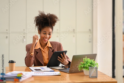 Happy African American businesswoman received good news, celebrating success with arms up in front of laptop at office desk