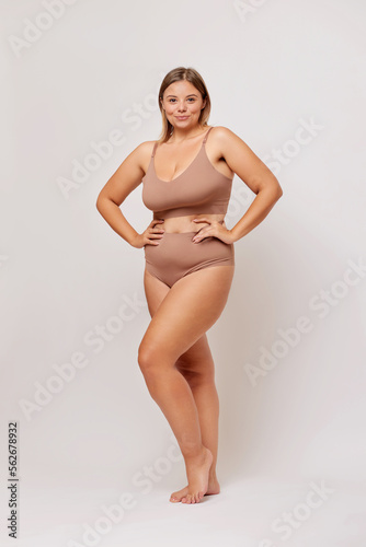 Vertical shot of cheerful young woman poses in beige lingerie, feels comfortable in her body, poses in studio against white background. 