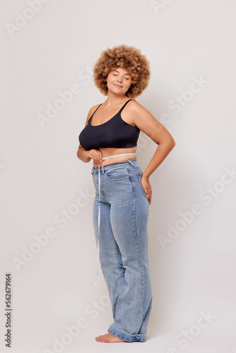 Vertical shot of overjoyed cute curly haired woman holds centimeter, checks her parameters and feels satisfied, isolated next to white wall. Concept of body positive, beauty, fashion, style