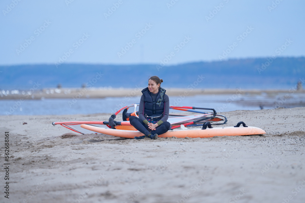 A mature woman, a windsurfer, is waiting on the sandy shore for other surfers on a cloudy autumn day. A mature woman, a windsurfer, is waiting on the sandy shore for other surfers on a cloudy autumn day.