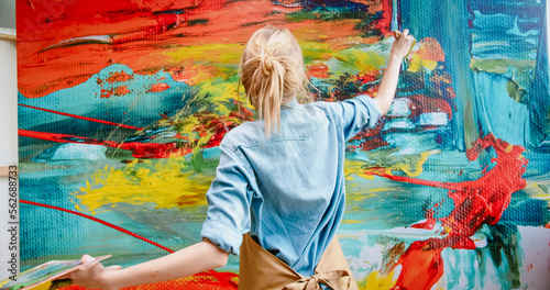 Close up of Caucasian cheerful happy young female artist in apron working in art studio painting using paint brush on big canvas making emotional colorful abstract masterpiece. Modern art concept