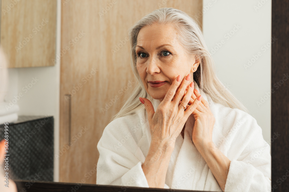 Senior woman with long grey hair touching her cheek with fingers in bathroom - obrazy, fototapety, plakaty 