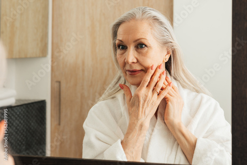 Fototapeta Senior woman with long grey hair touching her cheek with fingers in bathroom