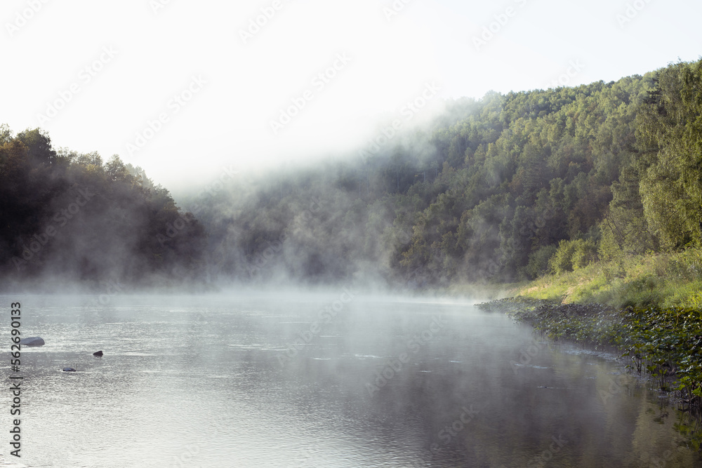 Calm summer morning sunrise landscape on river - smooth cold white haze steaming above water, lush green forest on slopes in bright sunlight. Mysterious and sinister scene in wild nature.
