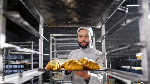 Caucasian handsome man in hat and uniform working in bakehouse and putting croissants on shelf. Male baker making fresh pastries in kitchen of bakeshop in morning. Confectionery producing concept.
