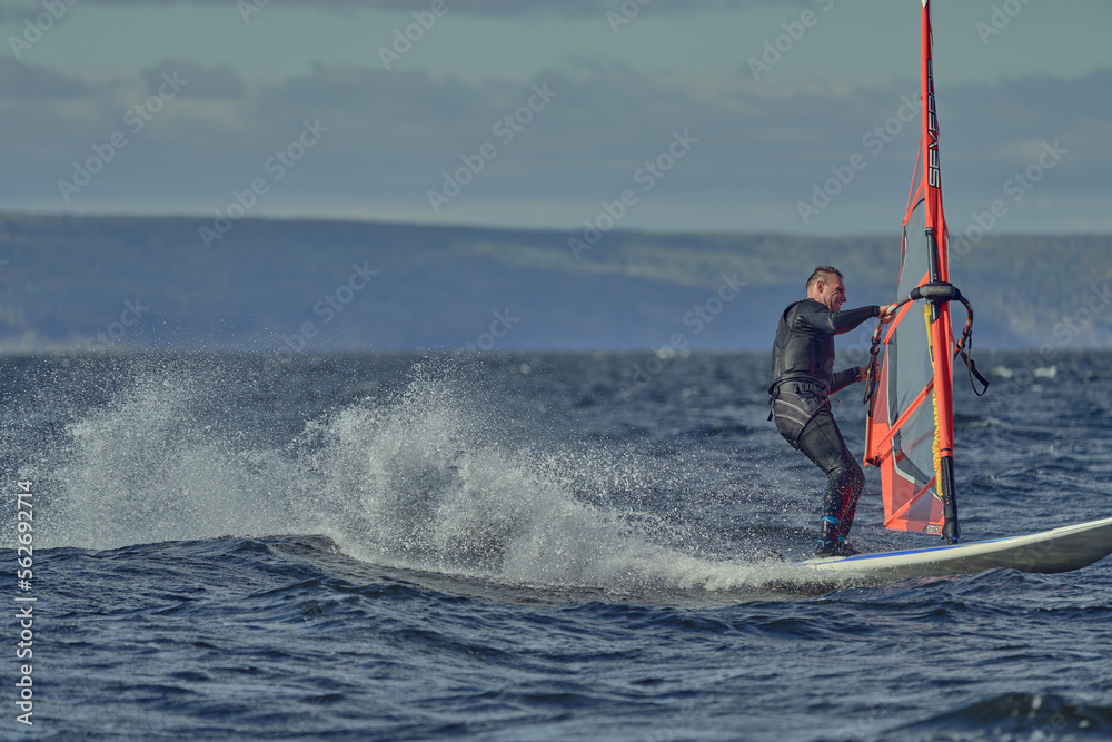 Obraz premium A mature man, an experienced windsurfer, rushes along the surface of the water, splashes scatter to the sides, a sunny autumn day.