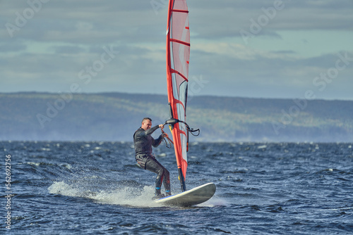 A mature man, an experienced windsurfer, rushes along the surface of the water, splashes scatter to the sides, a sunny autumn day.