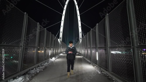 Wide shot of a man texting and walking on a bridge at night in the city