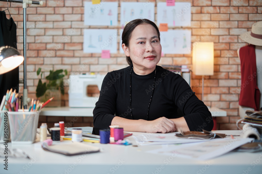 Asian mature female fashion designer is sitting at table, looking at camera and in studio, working with creative and sewing for dress design collection, professional boutique tailor SME entrepreneur. Asian mature female fashion designer is sitting at table, looking at camera and in studio, working with creative and sewing for dress design collection, professional boutique tailor SME entrepreneur.