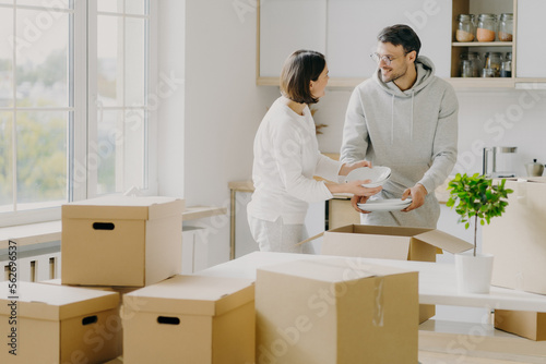 Photo of busy family couple unpack personal stuff from carton boxes, dressed in casual clothes, hold white plates, pose in spacious kitchen with modern furniture, surrounded with pile of packages