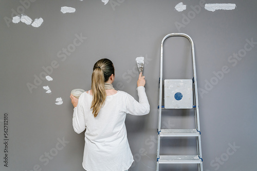 Backwards Blonde woman plastering with a spatula a wall after being painted with a grey painting. The painter is next to her ladder while covering small holes on the wall. Horizont photo