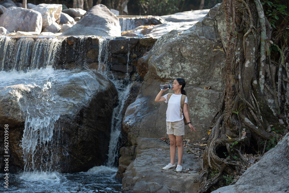 Female hiker looking at waterfall. Happy teenager backpacker enjoying amazing tropical waterfall raised hands travel lifestyle,  Female hiker crossing the forest creek. Young woman drinks water.
