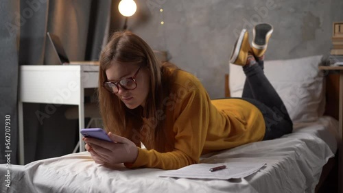 teenager girl student in glasses lies on a bed in a college dormitory holding a smartphone in her hands and communicates with friends online in social networks, writes and reads a message. photo