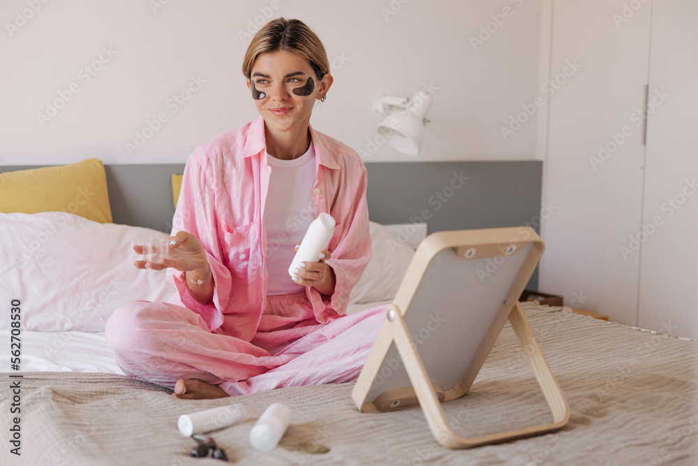 Pleasant young caucasian woman with black eyes patches holds face skin care product sitting on bed. Blonde hair female dressed in pink pajamas indoor. Concept organic cosmetics.