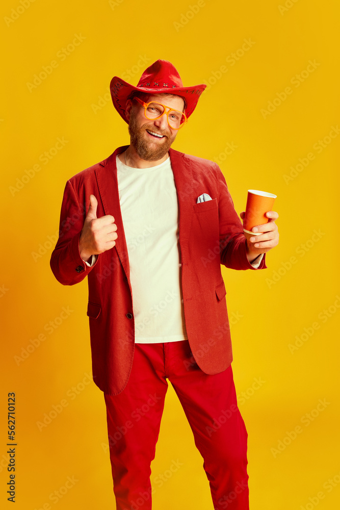 Fashionable man, hipster wearing bright red costume and cowboy hat posing over yellow background. Concept of style , fashion, happy mood, positive emotions Fashionable man, hipster wearing bright red costume and cowboy hat posing over yellow background. Concept of style , fashion, happy mood, positive emotions