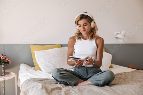 Calm young caucasian girl wears casual clothes sitting on bed and inspired takes notes in notebook. Cute girl with headphones spending time at home. Concept inspiration.