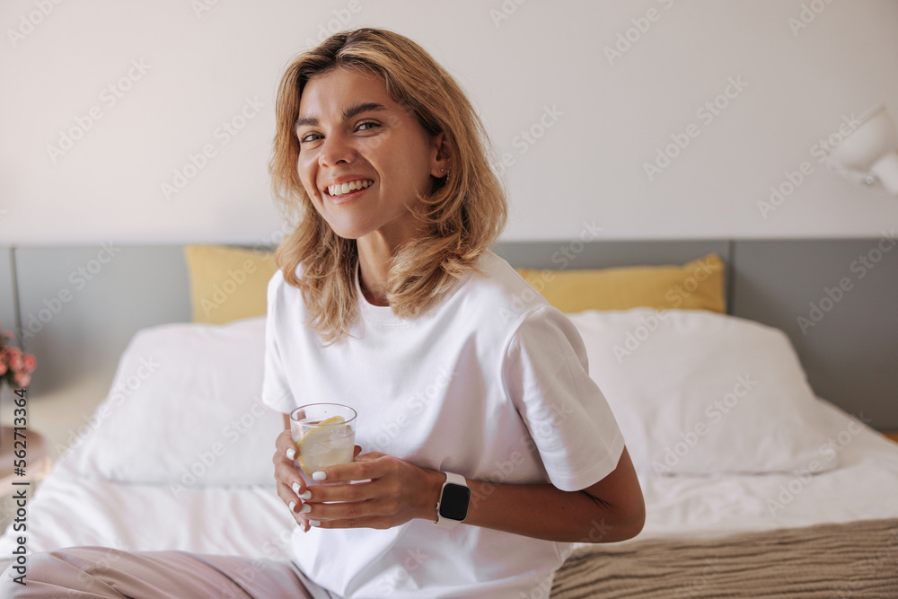 Happy young caucasian woman in pajamas holds glass of water with slice of lemon. Smiling girl is sitting on bed in her bedroom morning. Healthy morning concept. Happy young caucasian woman in pajamas holds glass of water with slice of lemon. Smiling girl is sitting on bed in her bedroom morning. Healthy morning concept.
