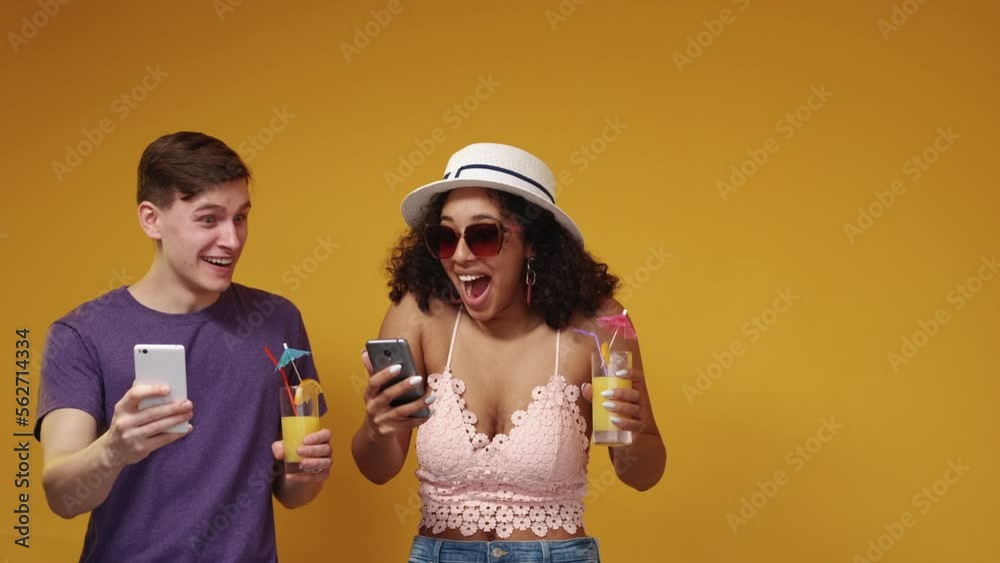Hot tours. Surprised couple. Advertising background. Excited man and woman getting unexpected information in smartphone posing with cocktails on yellow copy space. Hot tours. Surprised couple. Advertising background. Excited man and woman getting unexpected information in smartphone posing with cocktails on yellow copy space.