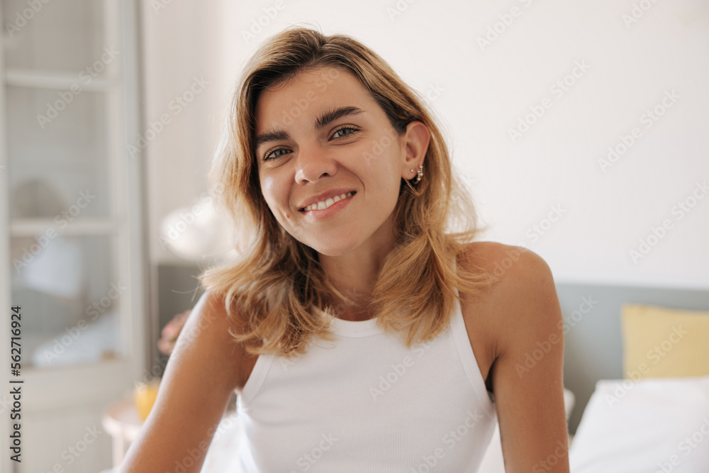 Close-up cheerful young caucasian girl wear white t-shirt looking at camera with smile indoor. Woman blonde hair posing on bed in bedroom. Concept portrait, beauty, lifestyle. Close-up cheerful young caucasian girl wear white t-shirt looking at camera with smile indoor. Woman blonde hair posing on bed in bedroom. Concept portrait, beauty, lifestyle.