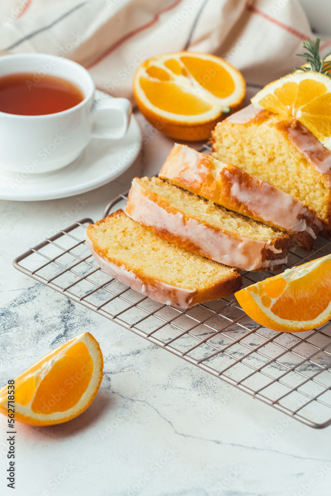 Loaf of orange bread covered with a confectionery glaze with lemon juice and decorated with orange slices. Chiffon cake on a pastry grill next to cup of tea. White background