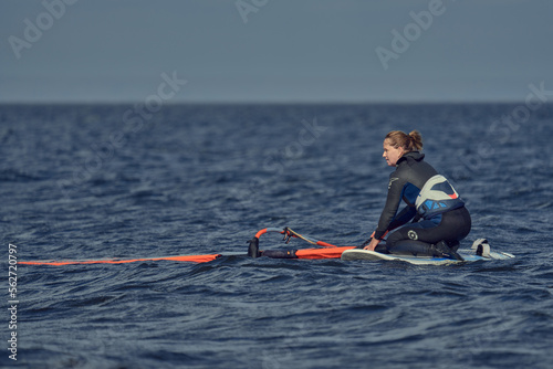 Mature woman, beginner windsurfer, sits on a sailboard floating in the water. Break from surfing on a sunny autumn day.