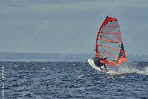A man in a wetsuit, a windsurfer, quickly glides through the waves on a sailboard on an autumn sunny day.