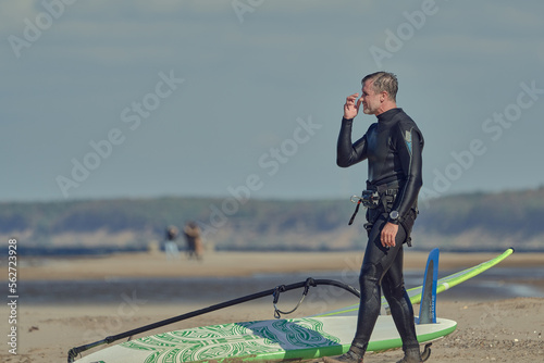 A mature man in a wetsuit, a windsurfer, stands on a sandy shore near a sailboard and rests.