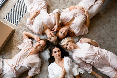 Above view of beautiful bride with veil, wearing in white nightgown, lying on floor and surrounded by pensive girls in pink peignoir, which together posing and looking at camera during bridal morning
