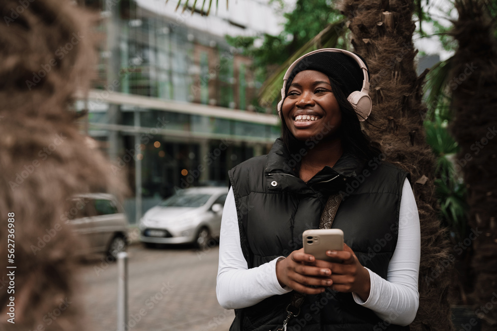 Fototapeta premium Smiling afro woman using mobile phone while standing outdoors at city street