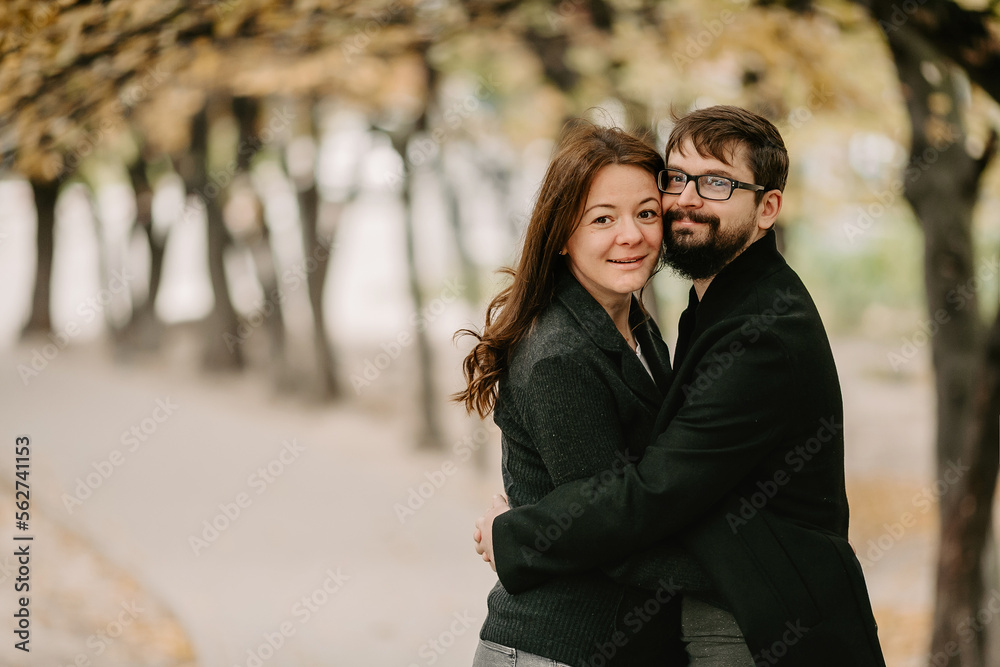 Portrait of loving couple cuddling on alley during autumn weather. Happy man with beard and eyeglasses, hugging with his girlfriend, while standing in park with yellowed leaves and looking at camera