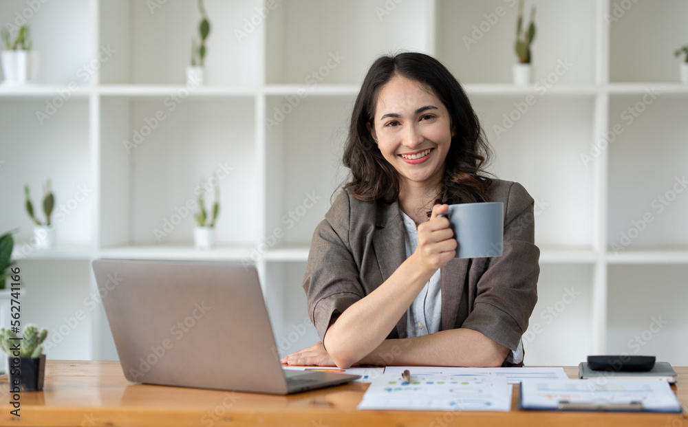 Asian businesswoman holding a coffee mug with a smile and working on a laptop computer with chart data The graph shows the results of the real estate business finance. Asian businesswoman holding a coffee mug with a smile and working on a laptop computer with chart data The graph shows the results of the real estate business finance.