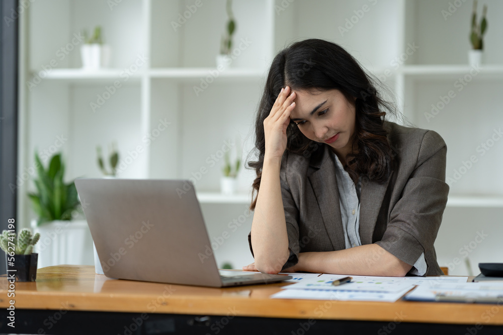 Tired stressed Asian businesswoman working on a laptop while sitting at a desk at the office and holding hands on temples. Migraine symptoms while calculating business income profit margin. Tired stressed Asian businesswoman working on a laptop while sitting at a desk at the office and holding hands on temples. Migraine symptoms while calculating business income profit margin.