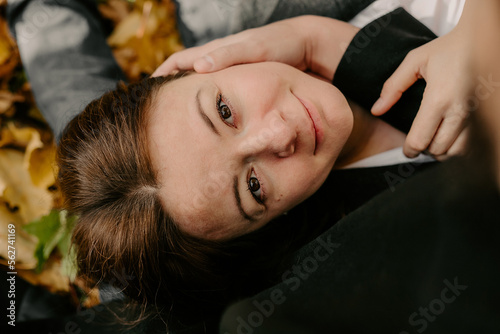 Portrait of attractive woman lying on yellowed leaves and looking at camera. Girl with dark hair and brown eyes, lying on legs of her boyfriend during romantic picnic in autumn park