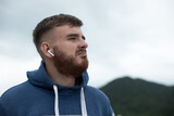 Portrait of bearded handsome pensive thoughtful guy, listen music in earphones, young upset sad frustrated man on natural mountain background, thinking with a serious face