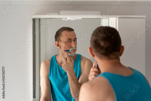 Wallpaper Mural Young attractive man removes his beard with razor with shaving foam on face. Reflection in mirror. Face care Torontodigital.ca