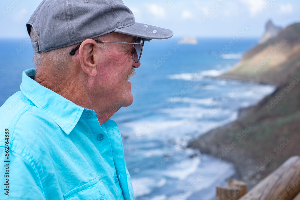 Portrait of old senior man with cap and glasses enjoying beauty in nature in Tenerife, Canary islands looking at sea. Happy retirement or vacation and free lifestyle