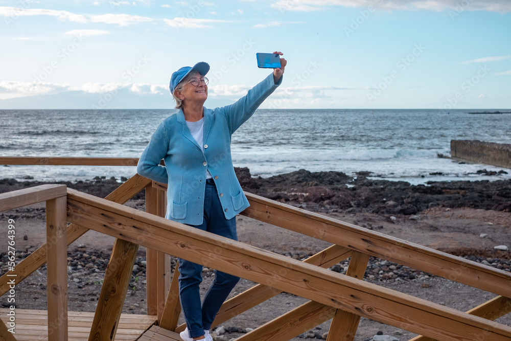 Carefree senior woman in hat and glasses standing outdoors at sunrise making a selfie with sea view. Smiling elderly lady holds the phone in her hand enjoying the silence of the morning on the beach