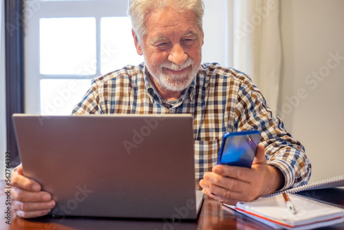Smiling mature bearded man sitting at table with laptop and books following online course. Senior man in checkered shirt enjoying teaching activity and learning using new technology