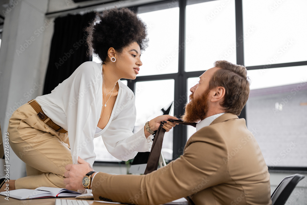 Fototapeta premium side view of passionate african american woman sitting on desk and pulling tie of bearded coworker in office.