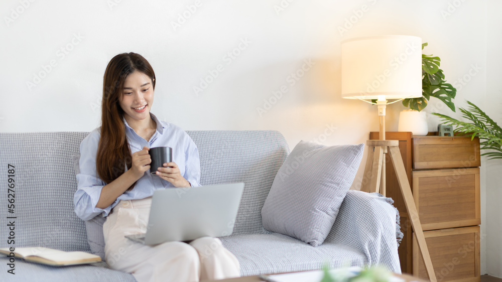 Beautiful Asian woman working on laptop and sipping coffee with smiling face in her home, Creating happiness at work with a smile, Freelancer working at home happily, Work from home. Beautiful Asian woman working on laptop and sipping coffee with smiling face in her home, Creating happiness at work with a smile, Freelancer working at home happily, Work from home.