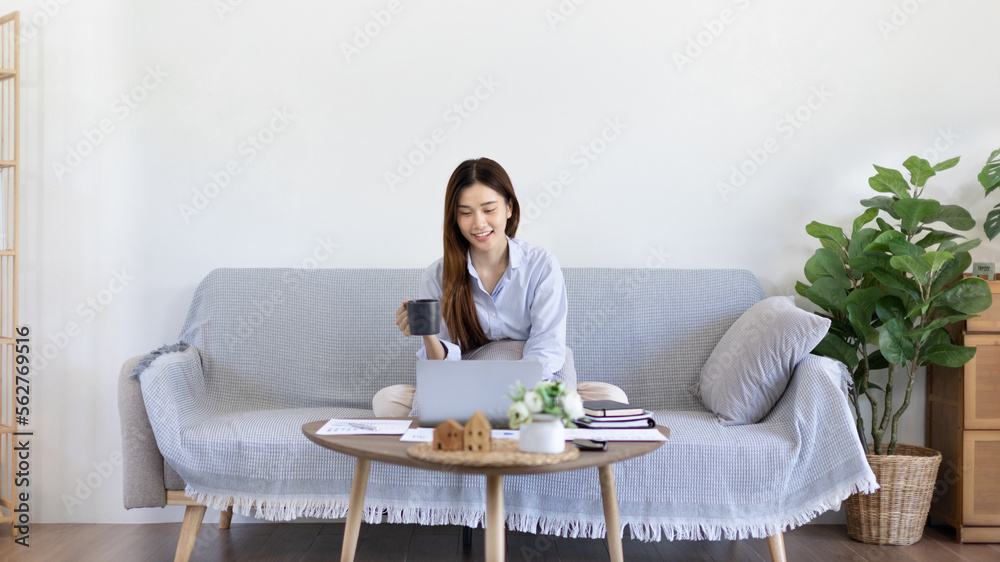 Beautiful Asian woman working on laptop and sipping coffee with smiling face in her home, Creating happiness at work with a smile, Freelancer working at home happily, Work from home. Beautiful Asian woman working on laptop and sipping coffee with smiling face in her home, Creating happiness at work with a smile, Freelancer working at home happily, Work from home.